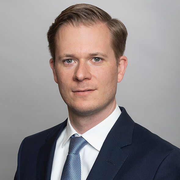 A professional headshot of a man in a dark suit with a white shirt and a patterned tie. He has short, light-colored hair and is looking directly at the camera with a neutral expression against a light gray background.