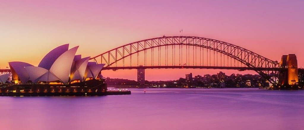 Das Bild zeigt das Sydney Opera House mit seinen charakteristischen, segelartige Dächern und die Sydney Harbour Bridge, die über den ruhig glühenden Hafen spannt. Der Himmel hat einen sanften, lila Farbton während der Dämmerung.