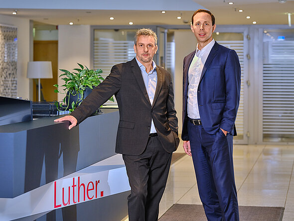 Two professional men stand beside a reception desk in a modern office setting. One wears a dark suit with a light shirt, while the other is dressed in a blue suit. In the background, there is a plant and subtle decorative elements. The company name "Luther." is visible on the desk.
